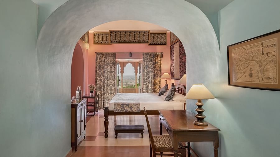 A view through an arched doorway of a room with a bed, a desk, and a framed artwork in Tijara Fort-Palace - 19th Century, Alwar.