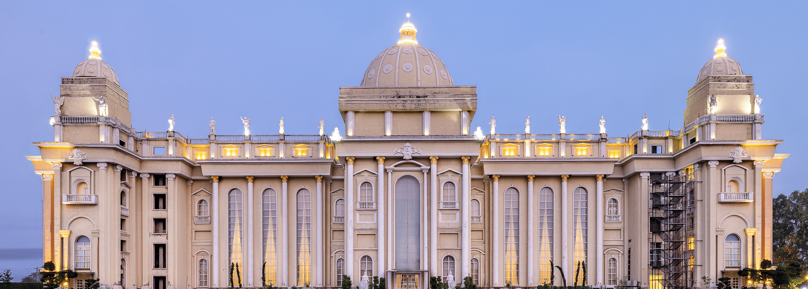 Vast, ornate, pale-coloured building with a central dome and grand architecture, illuminated against a twilight sky, set on a lawn at Hotel Hukam's Lalit Mahal.