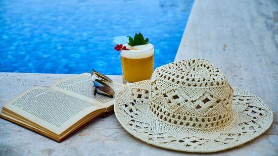 A glass of juice and a hat placed on a poolside lounge chair next to the swimming pool, with the water shimmering in the background.