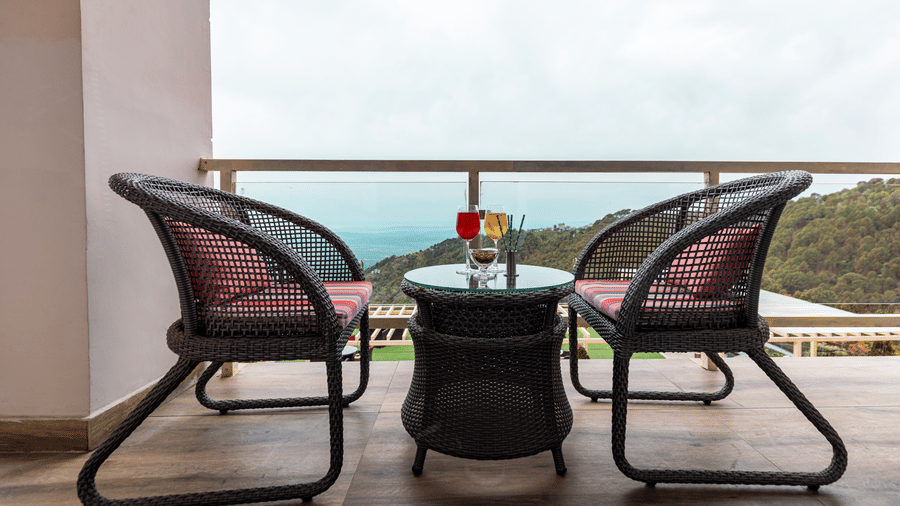 A balcony with 2 chairs and a table placed facing an open view with hills in the distance at Indraprastha Resort & Spa, McLeod Ganj.