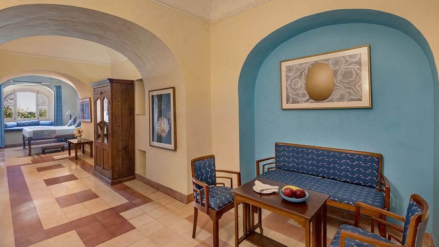 A room with a sofa, two chairs, a table, and framed artworks, with an arched doorway leading to another room in Tijara Fort-Palace - 19th Century, Alwar.