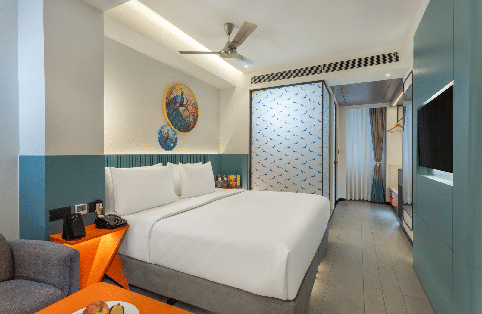 A bedroom featuring a neatly made bed, a wall-mounted television screen facing the bed,  a separate sitting area, and a ceiling fan | Zibe Luxe Hyderabad by GRT Hotels  