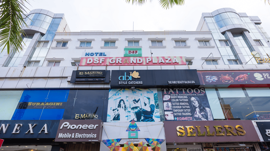 The daytime exterior of DSF Grand Plaza, Tuticorin showing the Hotel DSF Grand Plaza signage above ground-floor shopfronts, framed by overhanging palm fronds.