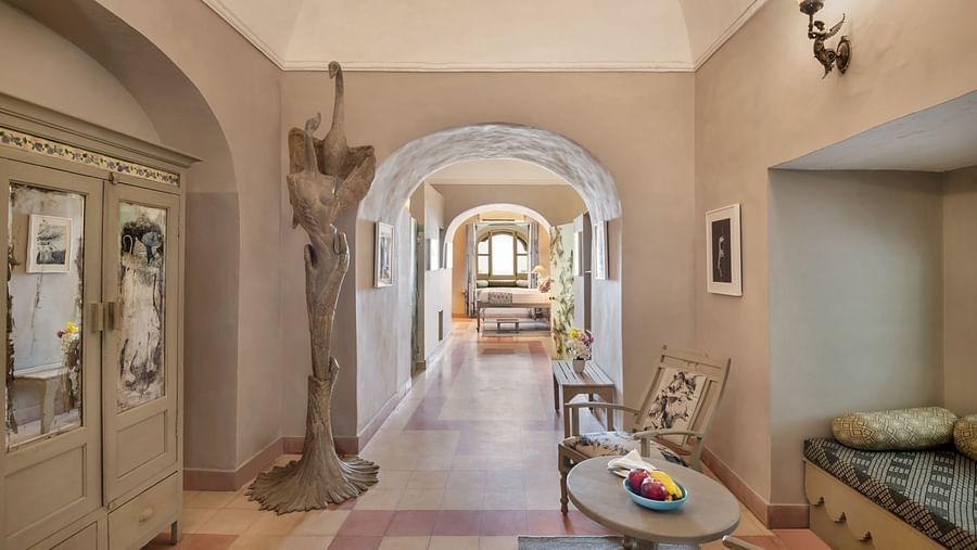 A hallway with arched doorways leading to other rooms, featuring a decorative cabinet, a sculpture, a table with chairs, and a built-in cushioned seating area in Tijara Fort-Palace - 19th Century, Alwar.