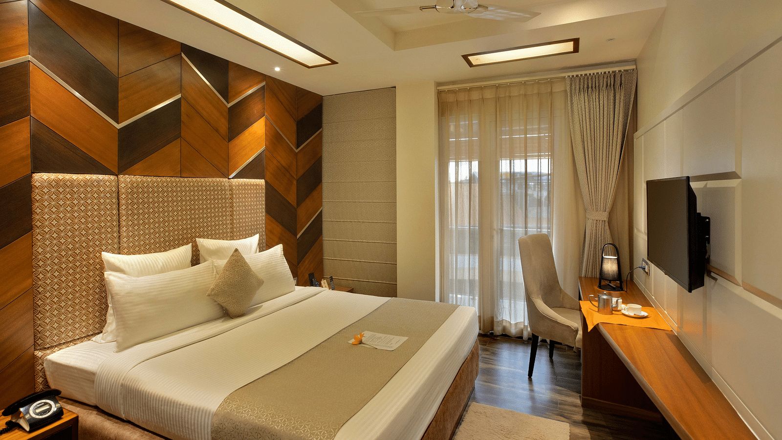Image of warmly lit Coral Suite at Coral Reef Hotel and Spa in Port Blair featuring a plush bed in front of a TV mounted on the wall and next to it is a work desk and a chair near the door to balcony