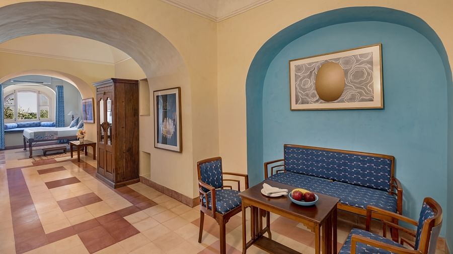 A room with a sofa, two chairs, a table, and framed artworks, with an arched doorway leading to another room in Tijara Fort-Palace - 19th Century, Alwar.
