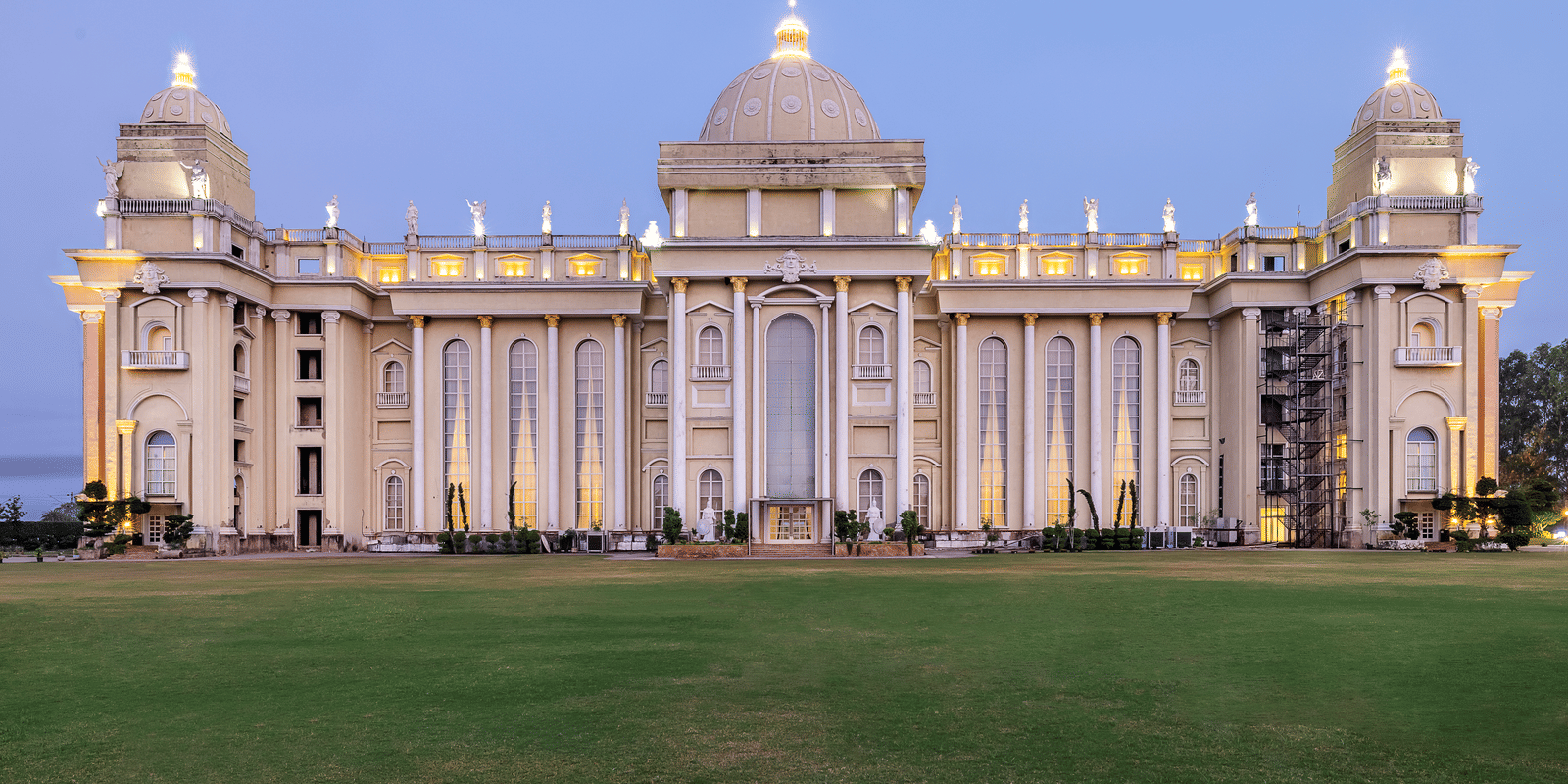 Vast, ornate, pale-coloured building with a central dome and grand architecture, illuminated against a twilight sky, set on a lawn at Hotel Hukam's Lalit Mahal.