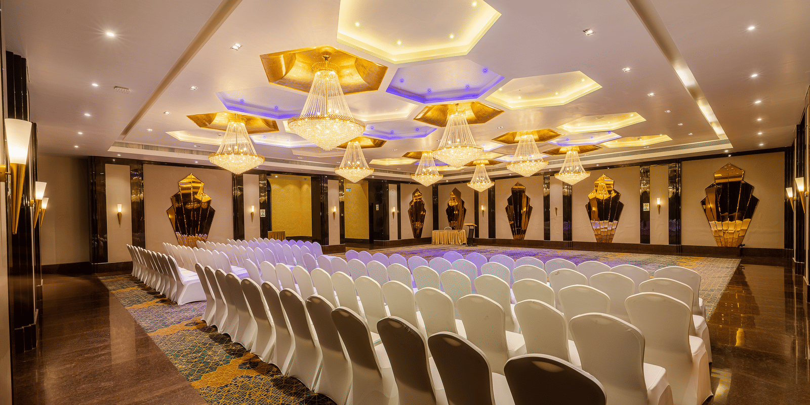 Banquet hall set with rows of white chairs facing a stage area, brightly lit by impressive hexagonal ceiling fixtures at Hotel Hukam's Lalit Mahal.