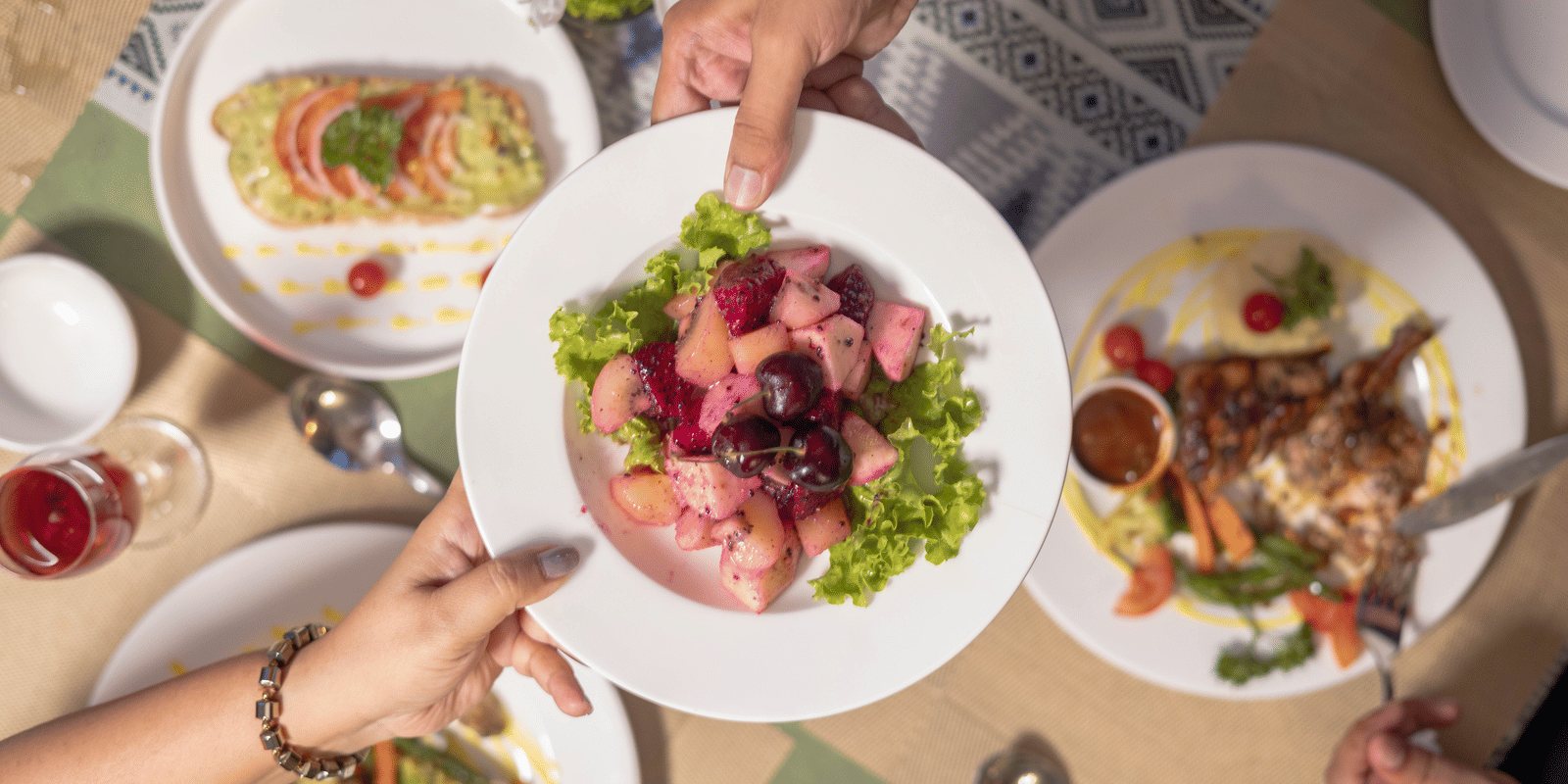 Plates of salad and dishes on a dining table featuring a dish at the middle held by two hands on the either side at Megha Resort & Villas, Dehradun.