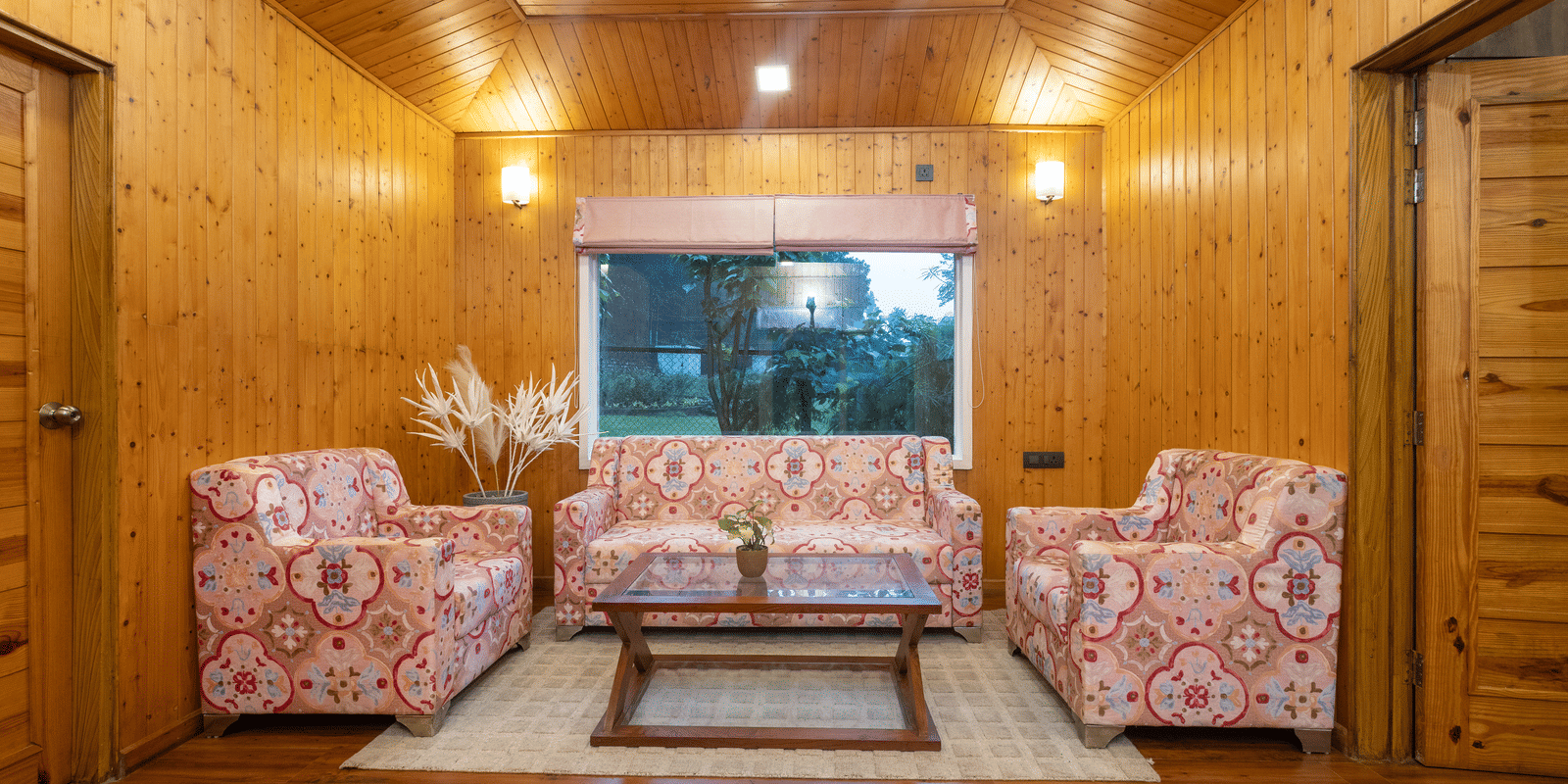 A cosy living room featuring floral-patterned sofas, wooden walls, and a view through a wide window at Megha Resort & Villas, Dehradun.