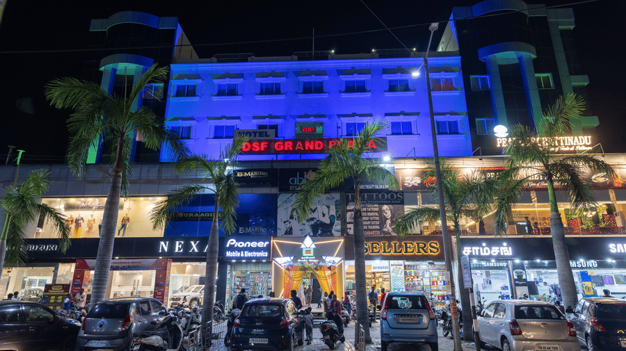 Night-time exterior of DSF Grand Plaza, Tuticorin illuminated in blue, with the hotel signage above ground-floor shops, palm trees, and parked cars outside.