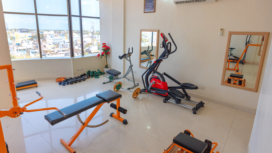 Hotel gym at DSF Grand Plaza, Tuticorin with a cross-trainer, weight bench, dumbbell rack, and wall mirrors, with windows overlooking the city.
