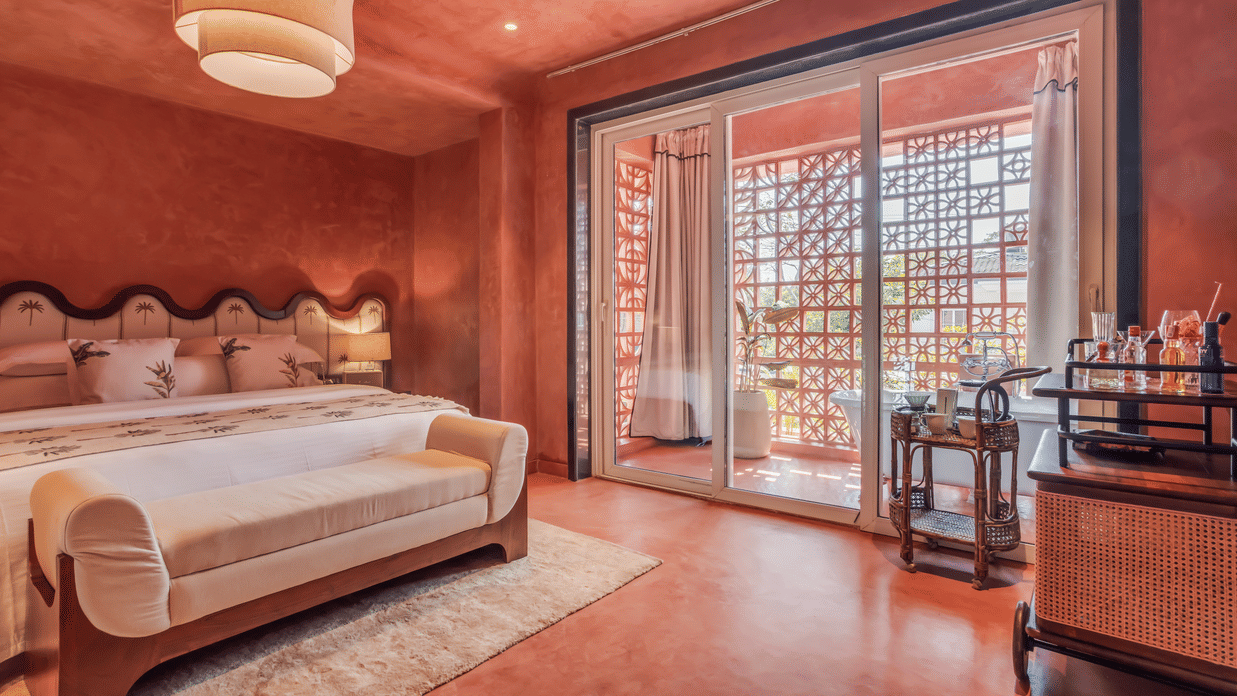 Bedroom at Unseen Boutique Hotel with reddish?brown walls, tufted headboard, white rug, sliding glass door to balcony, and side cart with bottles and glasses.