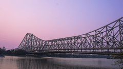 Howrah Bridge at dusk against a soft pink sky, its silhouette stretching across the river in a calm, reflective setting