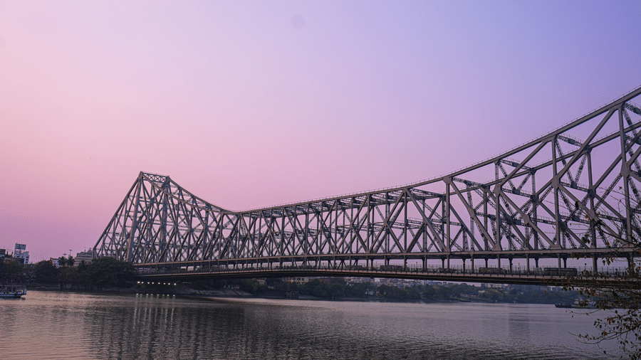 Howrah Bridge at dusk against a soft pink sky, its silhouette stretching across the river in a calm, reflective setting