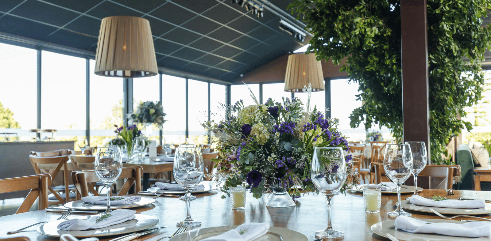 A well-appointed restaurant setting with tables set with fine china and glassware, surrounded by natural light and greenery.