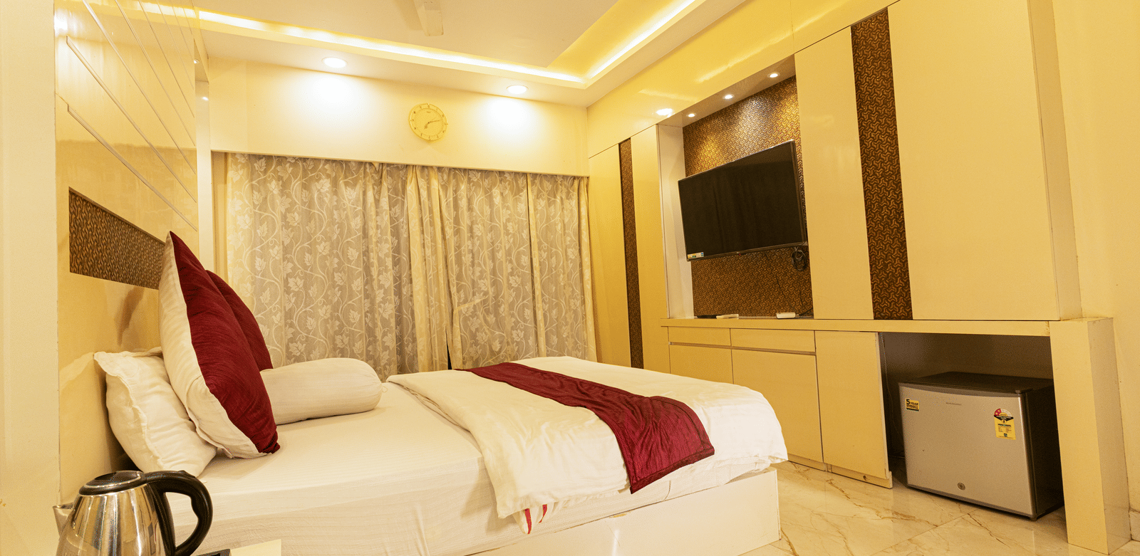 A bedroom with a king size bed, a bedside table with an electric kettle, a television, a mini fridge, and curtains at Hotel Seagull Digha.