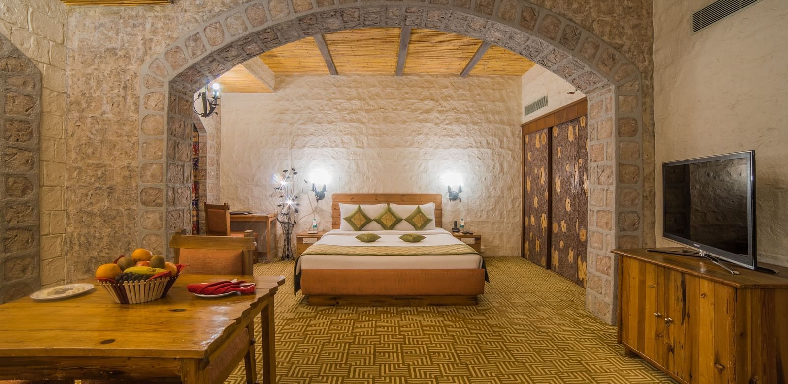 A spacious hotel bedroom with stone walls, wooden furniture, and a welcoming bed under a vaulted ceiling at  Sitara Luxury Hotel in Hyderabad near Ramoji Film City.