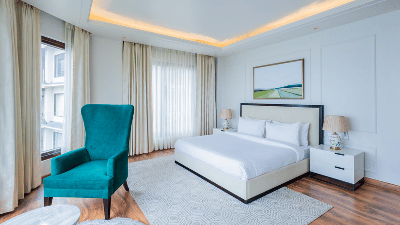 Bright, airy bedroom decorated in white tones with accent furniture and expansive windows, reflecting the stylish and contemporary interiors found at The Manor Sports & Wellness Hotel in Shimla.
