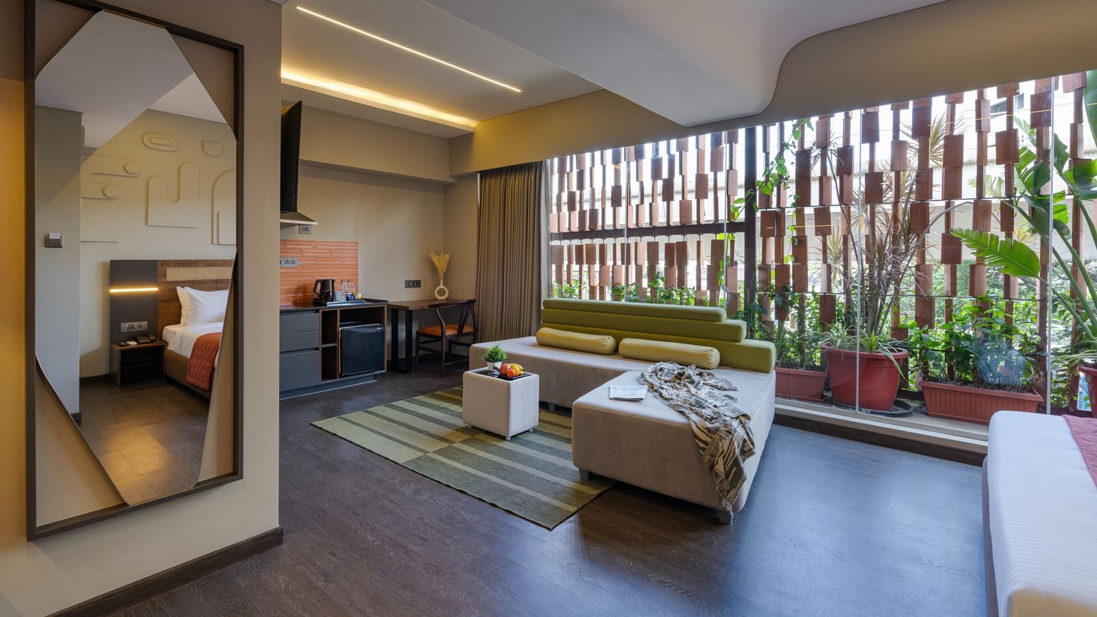 A lounger sofa with with a dining table on one end and open space in the other end, behind it the wall has transparent glass and a lot of plants can be seen through it.