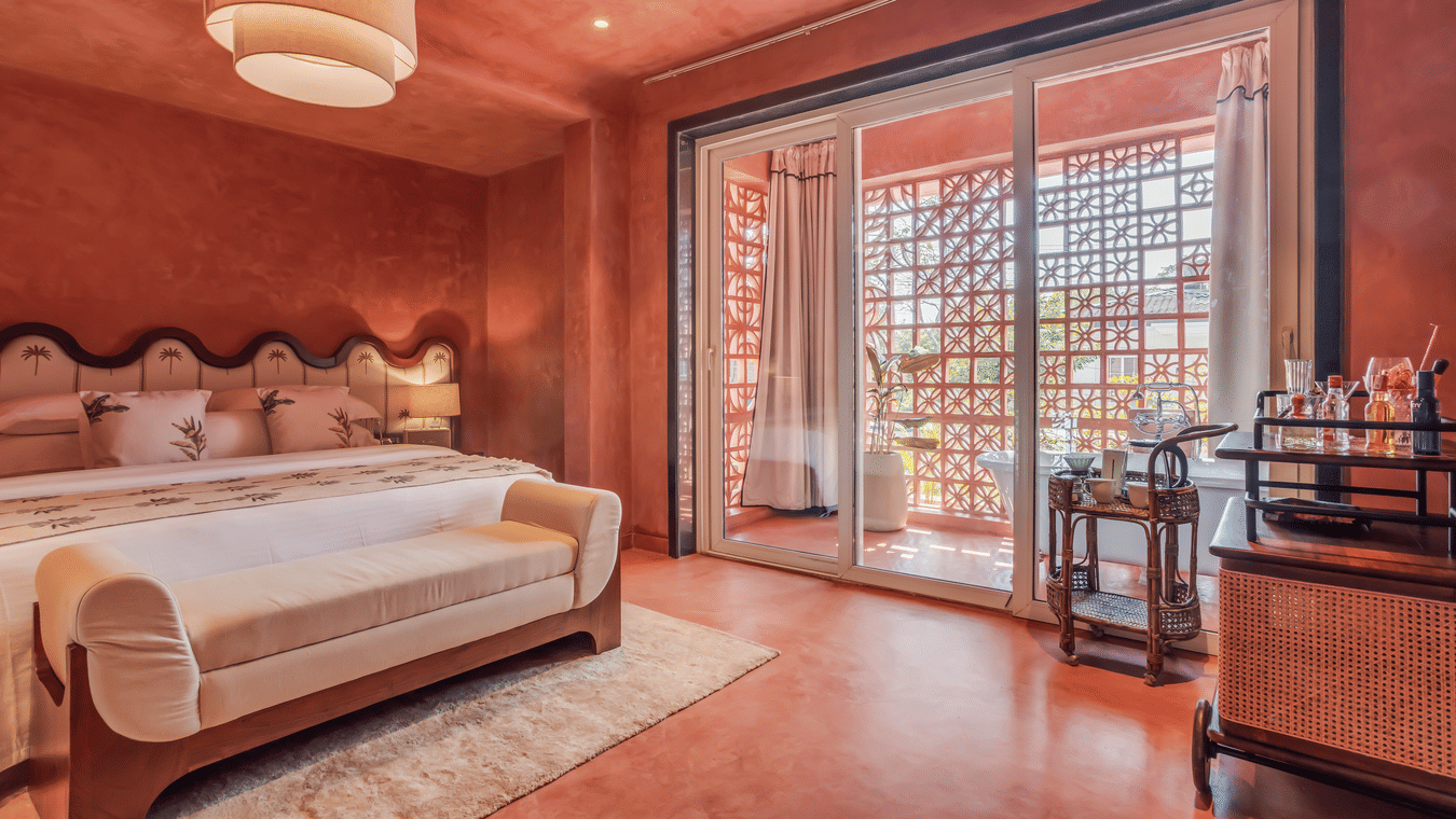 Bedroom at Unseen Boutique Hotel with reddish?brown walls, tufted headboard, white rug, sliding glass door to balcony, and side cart with bottles and glasses.