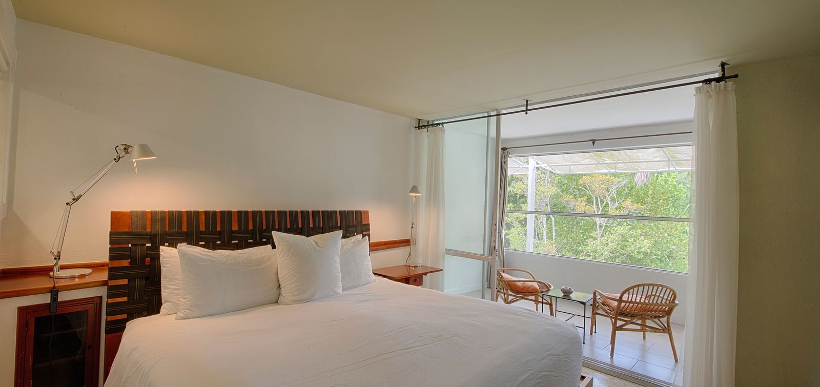 Bed with white linens at Casa Morada, facing a window with a garden view and two chairs.