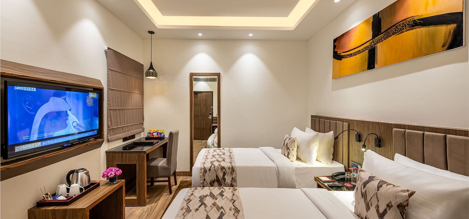 A hotel room with two twin beds, a flat-screen TV, and a work desk. It features a unique tray ceiling with lighting, a ceiling fan, and colourful abstract artwork - Lords Eco Inn Dehradun. 