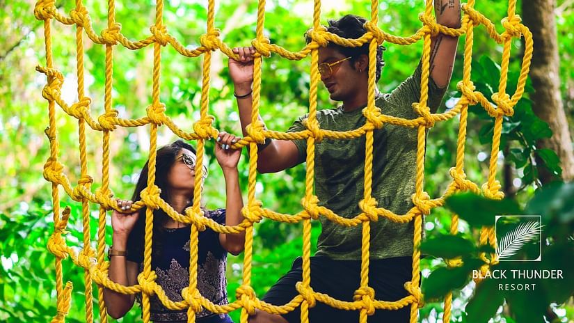 Two people participating in a rope obstacle course in a forested area, holding onto a vertical rope net - Black Thunder, Coimbatore