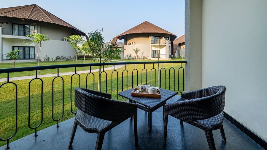 An image of a balcony with two wooden chairs and a tea table from where other villas and lawn can be seen