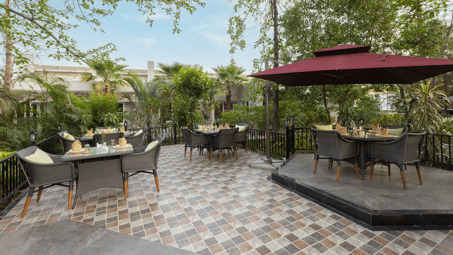 An outdoor dining area with tables set under a large red umbrella and trees, featuring a tiled floor  at The Golden Tusk, Jim Corbett