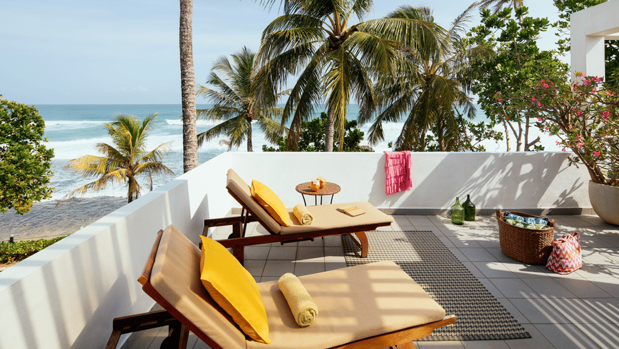 A view of 2 wooden lounge chairs at Owl and the Pussycat Hotel in Galle with head-rest cushions at an open balcony overlooking a beach lined with coconut trees and crashing ocean waves.