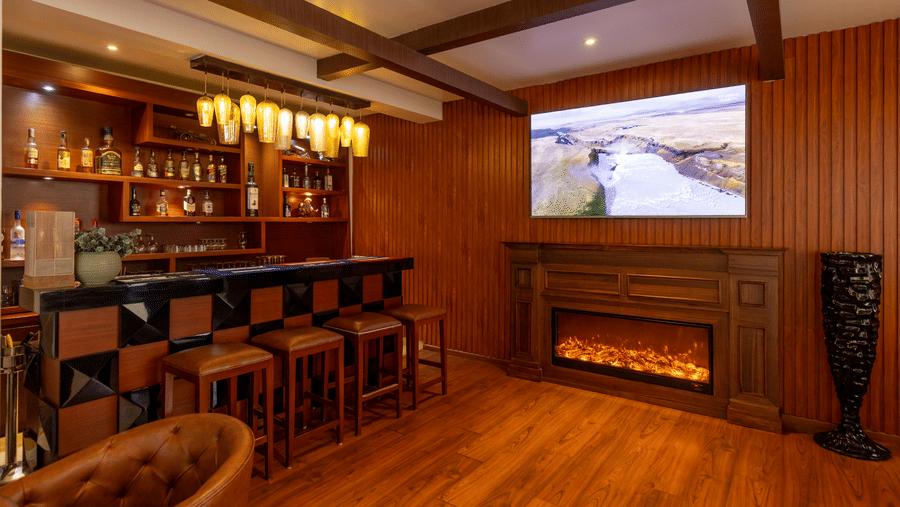 A bar with multiple stools, a counter, shelves with bottles, and a screen mounted on the wall at Indraprastha Resort & Spa, McLeod Ganj.