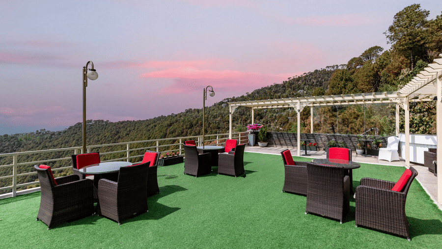 An outdoor seating area with tables, chairs, and a view of hills during evening at Indraprastha Resort & Spa, McLeod Ganj.
