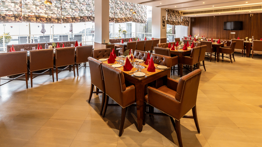A restaurant with tables, chairs, and large windows allowing light inside at Indraprastha Resort & Spa, McLeod Ganj.