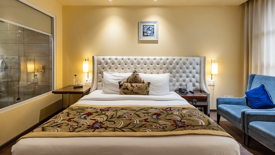 A bedroom with a king size bed, side tables, lamps, sofa, and a glass partition visible at Indraprastha Resort & Spa, McLeod Ganj.