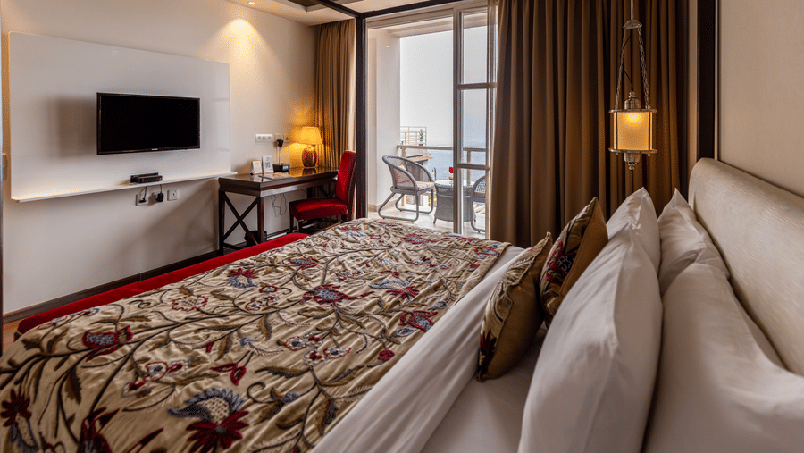 A bedroom with a king size bed, wall-mounted TV unit, side tables, lamps, work desk, and curtains arranged indoors at Indraprastha Resort & Spa, McLeod Ganj.