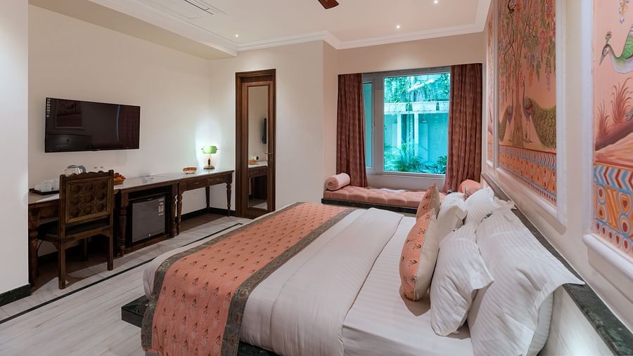 A headboard view of a spacious bedroom at Panna Vilas Palace, displaying a wall-mounted television with a wooden table underneath and large windows inviting natural light