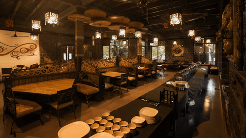 A wide view of dining area of Adamo The Village featuring rustic decor, ceiling lights, and wooden furniture arranged neatly in rows.