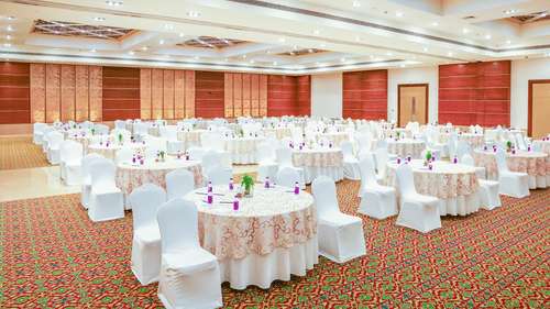 A spacious conference hall with numerous round tables set up, each covered with a white tablecloth and featuring a centrepiece - Balaji Sarovar Premiere, Solapur