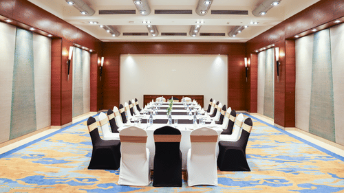 A modern meeting room featuring a long rectangular table surrounded by chairs, each covered with a gold chair cover, set up for a business event or conference - Balaji Sarovar Premiere, Solapur