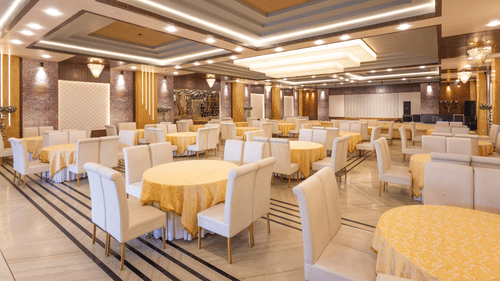 A spacious banquet hall is decorated with elegant cream and gold tables and chairs, ready for an event - Pride Premier Dehradun