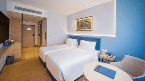A hotel bedroom with two beds and white linens is shown, with a large window and a painting on the wall | Standard Twin Room | Sarovar Portico, Sriperumbudur