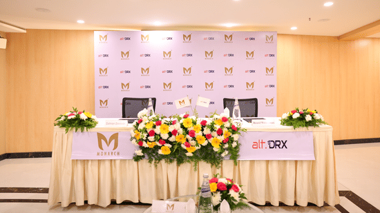 A banquet hall view of Coronet Ball Room at The Monarch Hotel, Brigade Road showing round dining tables with table linen covered chairs tied with sashes and a branded stage backdrop at the far end.