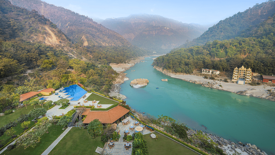 Riverside patio dining area overlooking a turquoise river, mountain landscape, landscaped lawns, and an outdoor pool beside the hotel.