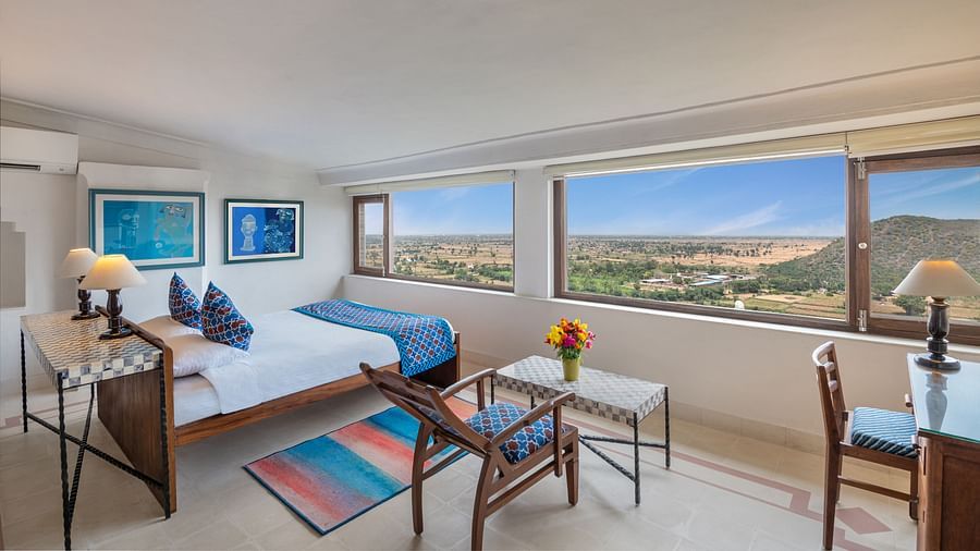 A room with a daybed, two framed artworks, a table with chairs, a desk, and large windows with a scenic view in Tijara Fort-Palace - 19th Century, Alwar.
