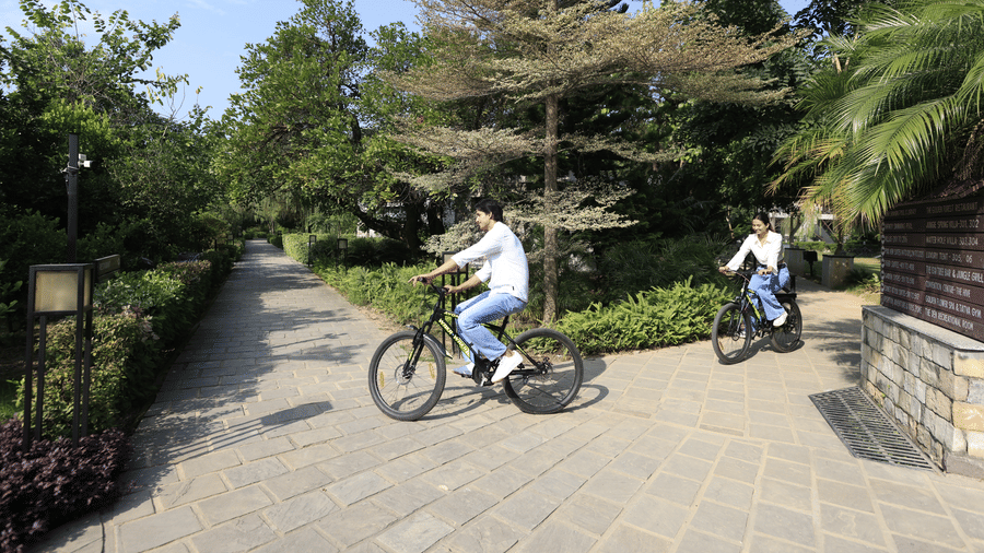 Two people riding bicycles on a paved path outdoors with trees and greenery at The Golden Tusk, Jim Corbett