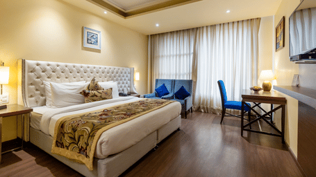 A spacious bedroom at Indraprastha Resort & Spa, McLeod Ganj, featuring a neatly-made bed, a table and a chair, and full-length curtains.