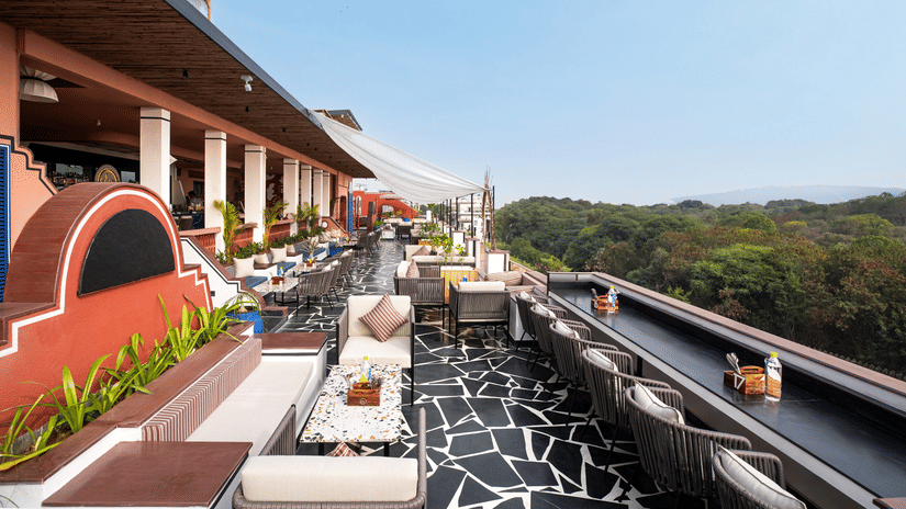 A rooftop terrace with chic outdoor furniture, a striking black and white floor, and lush greenery in the background at The Sierra, Udaipur.