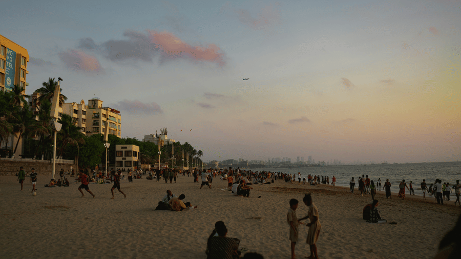Visitors relaxing, walking, and enjoying the lively evening atmosphere along Juhu Beach as the Mumbai skyline fades into the sunset sky.