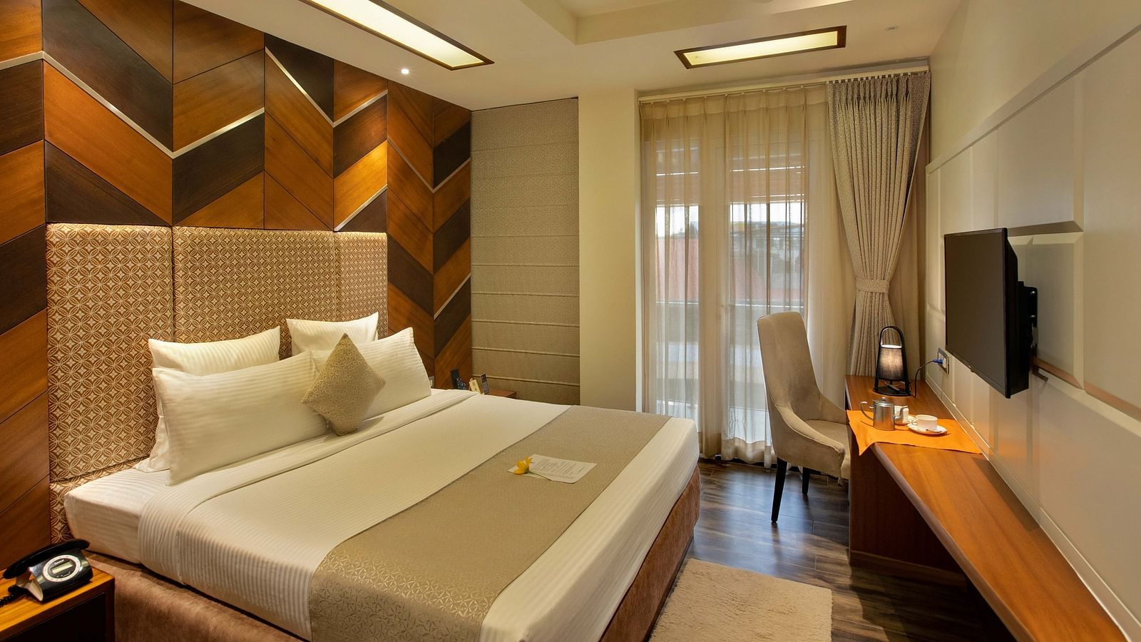 Image of warmly lit Coral Suite at Coral Reef Hotel and Spa in Port Blair featuring a plush bed in front of a TV mounted on the wall and next to it is a work desk and a chair near the door to balcony